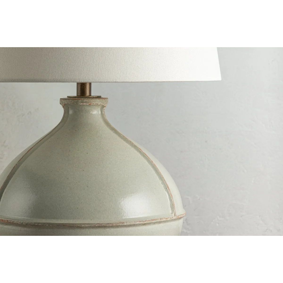 Inspired by rustic pottery, this table lamp's beautifully shaped base is crafted of pale sage ceramic giving it an earthy pale green tone. The off white linen shade pairs naturally and casts a soft glow. Raised seamlines and a reactive glaze that allows hints of the natural ceramic to peek through, give the fixture a hand-thrown, crafted feel. Amethyst Home provides interior design, new home construction design consulting, vintage area rugs, and lighting in the Austin metro area.